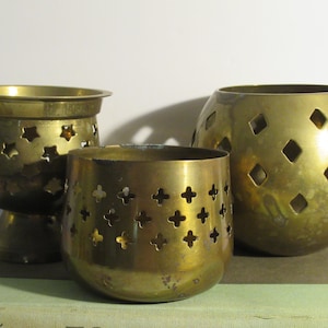 May include: Three brass candle holders with intricate cut-out designs. The holders are in different shapes and sizes, with one being round, one being a cylinder, and one being a pedestal.