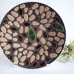 May include: A round decorative plate with a mosaic design of brown, pink, and green ceramic tiles. The tiles are shaped like acorns and leaves.