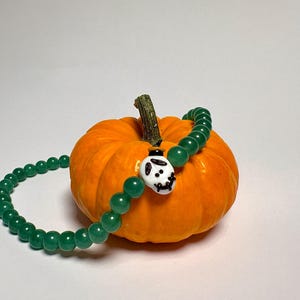May include: A small, round orange pumpkin with a green stem. A green beaded bracelet with a white and black skull charm is draped over the pumpkin. The bracelet is made of round, polished beads.