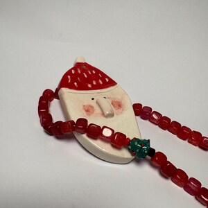 Puede incluir: Un collar con colgante de Papá Noel. El colgante es blanco con gorro rojo y mejillas rosadas. El collar presenta cuentas cuadradas rojas y una cuenta de acento verde y negro. Un accesorio festivo.