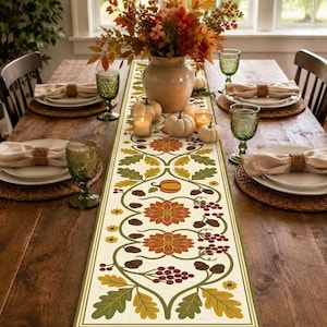 May include: A rectangular table runner featuring a cream background with a repeating pattern of orange flowers, green oak leaves, brown acorns, and small berries. The runner is centered on a wooden table with place settings and a floral centerpiece.