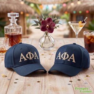 May include: Two navy blue baseball caps with the Greek letters AΦA embroidered in a light tan colour. The caps are on a wooden table with drinks and flowers in the background. The caps are the focal point of the image.