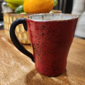 Pode incluir: Uma caneca de cerâmica vermelha com salpicos pretos e uma pega preta. A caneca tem uma borda branca. A caneca está sobre uma superfície de madeira. Provavelmente feita à mão.