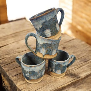 May include: Four handmade ceramic mugs stacked on a wooden surface. The mugs have a blue and white glaze with a speckled pattern and a natural, unglazed base. Each mug has a curved handle.