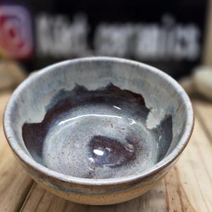 May include: A handmade ceramic bowl with a unique glaze. The interior features a swirl of dark brown and gray, while the rim has a light blue and white color. The bowl is set on a wooden surface, showcasing its handcrafted nature.