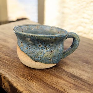 May include: A handmade ceramic mug with a rounded shape and a small handle. The mug features a speckled blue glaze on the upper portion, transitioning to a tan base. The mug is sitting on a wooden surface.