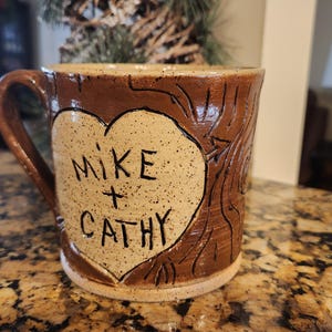 Puede incluir: Una taza de cerámica hecha a mano con exterior marrón y forma de corazón crema. El corazón está inscrito con "MIKE + CATHY" en letras negras. La taza tiene un diseño texturizado, tallado en madera.