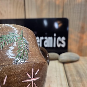 Può includere: Una tazza in ceramica marrone maculata con un design a ramo di pino verde intagliato a mano e accenti rosa a forma di stella. La superficie ha una finitura terrosa e testurizzata con dettagli intagliati visibili. La tazza è davanti a un cartello.