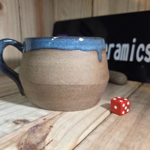 May include: A ceramic mug with a rounded body and a curved handle. The lower portion of the mug has a matte, light brown, textured finish, while the upper rim and handle feature a glossy, deep blue glaze that drips down the side. A small red die with white dots sits nearby.