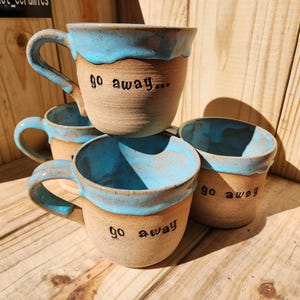 Op de afbeelding: Een stapel handgemaakte keramische mokken met een lichtbruine buitenkant en een turquoise blauwe binnenkant en rand. Elke mok heeft de tekst "go away..." gegraveerd.