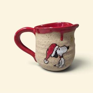 May include: A ceramic creamer pitcher with a speckled beige finish and a bright red glazed rim and handle. The body features a hand-painted illustration of a white beagle wearing a red Santa hat, with a small yellow star detail above the character.