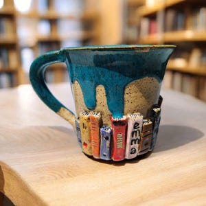 May include: A handmade ceramic mug with a teal glaze and a handle. The mug features a design of colorful books around the base, with the name "Emma" written on one of the books. The mug is sitting on a wooden surface.
