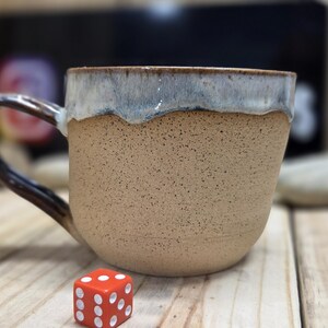 May include: A speckled beige ceramic mug with a dark brown handle and a light blue rim. A red die with white dots sits in front of the mug. The mug is on a wooden surface.