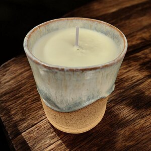 May include: A handmade candle in a ceramic container. The candle has a creamy white wax and a single wick. The container is a light blue and tan color, with a textured, organic design. The candle sits on a wooden surface.