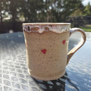 Pode incluir: Uma caneca de cerâmica artesanal com um corpo castanho salpicado e uma borda branca e castanha. A caneca apresenta três pequenos desenhos de corações vermelhos. A alça é curva. A caneca está numa mesa de vidro.