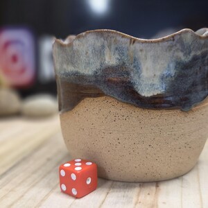 May include: A handmade ceramic pot with a wavy rim, featuring a speckled tan base and a dark blue and brown glazed top. A small red die with white dots rests beside the pot.