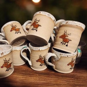May include: A collection of handmade ceramic mugs with a rustic, off-white glaze. Each mug features a hand-painted brown reindeer design with red accents. The mugs have a unique, organic shape and are stacked on a wooden surface.