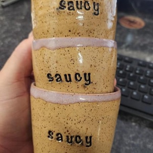 Könnte beinhalten: Drei gestapelte Keramikbecher mit gesprenkelter, hellbrauner Oberfläche und einem hellvioletten Rand. Auf jeder Tasse ist das Wort "saucy" in Schwarz aufgedruckt. Die Becher sind handgefertigt und haben eine rustikale Ästhetik.