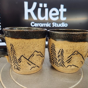 May include: Two ceramic mugs with a speckled tan base and dark brown rim and handle. The mugs feature a mountain and tree design in black. The text "Küet Ceramic Studio" is visible in the background.
