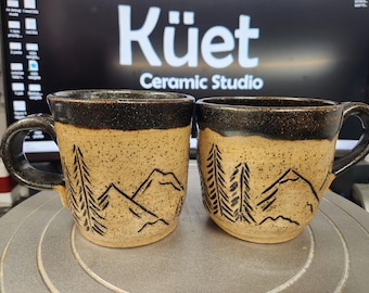 Könnte beinhalten: Zwei Keramikbecher mit gesprenkelter beigefarbener Basis und dunkelbraunem Rand und Griff. Die Becher haben ein Berg- und Baumdesign in Schwarz. Der Text "Küet Ceramic Studio" ist im Hintergrund sichtbar.
