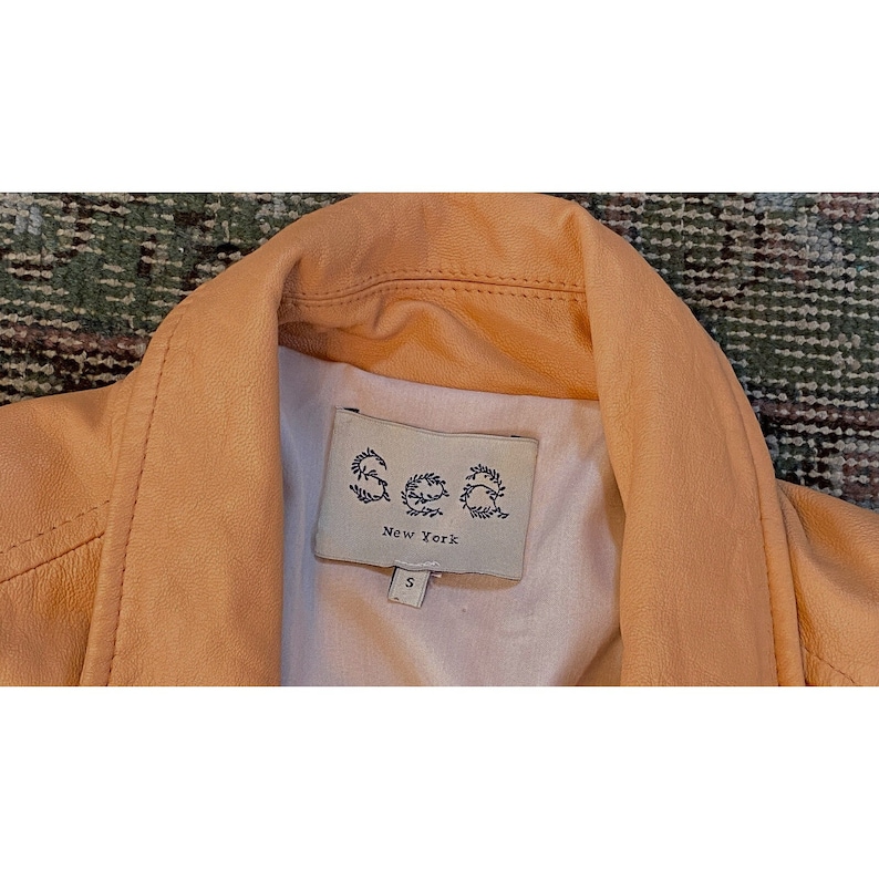 Pode incluir: Um close-up de uma jaqueta de couro bege com uma etiqueta que diz "Sea New York" e uma etiqueta de tamanho "S".