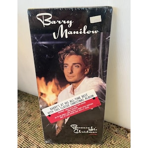 Puede incluir: Una cinta VHS sellada del álbum navideño de Barry Manilow, "Because It's Christmas". La portada presenta una foto de Manilow con una camisa blanca, con una chimenea en el fondo. Una etiqueta roja dice "Barry's at his all-time best with this classic Christmas album."