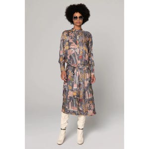Op de afbeelding: Een lange mouwen jurk met een kleurrijke bloemenprint en een strik in de taille. De jurk is donkerblauw met roze, gele en groene accenten. Het model draagt witte laarzen.