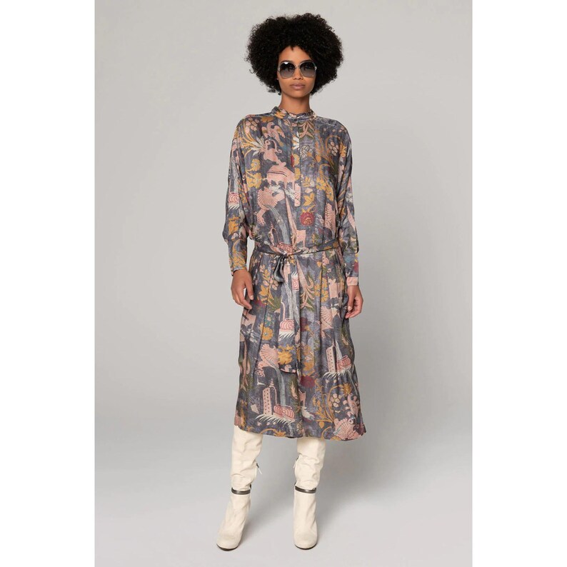 Op de afbeelding: Een lange mouwen jurk met een kleurrijke bloemenprint en een strik in de taille. De jurk is donkerblauw met roze, gele en groene accenten. Het model draagt witte laarzen.