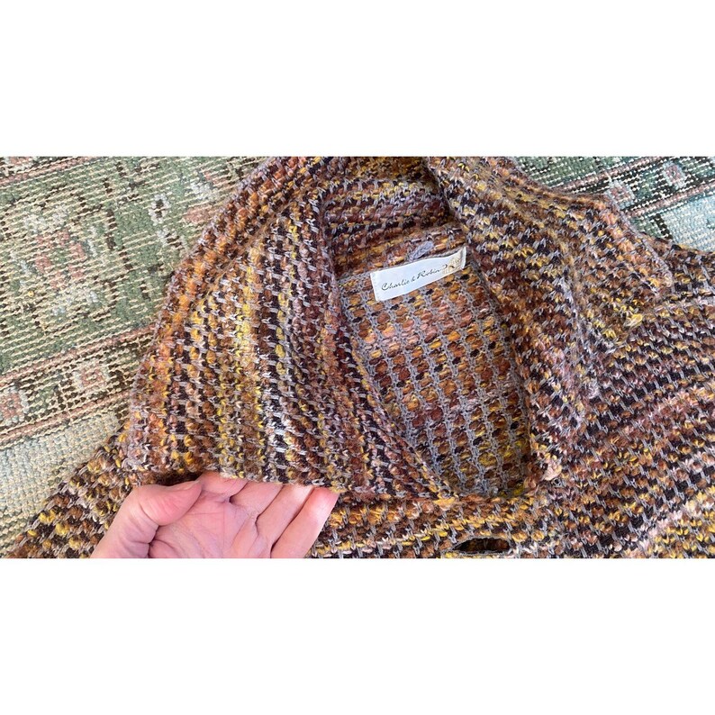 K&ouml;nnte beinhalten: Eine mehrfarbige Strickjacke in Braun-, Gelb- und Oranget&ouml;nen. Das Etikett lautet "Charles & Robin". Die Strickjacke hat einen Schalkragen und einen Knopfverschluss. Das Strickmuster ist strukturiert und scheint eine Mischung aus Wolle und anderen Fasern zu sein.