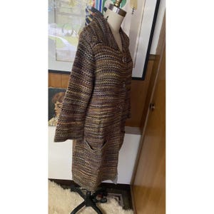 K&ouml;nnte beinhalten: Ein langer, mehrfarbiger Strickmantel mit Schalkragen und zwei Vordertaschen. Der Mantel wird auf einer Schaufensterpuppe pr&auml;sentiert und hat eine Knopfleiste vorne. Die Farbpalette umfasst Braunt&ouml;ne, Gelb und Schwarz.