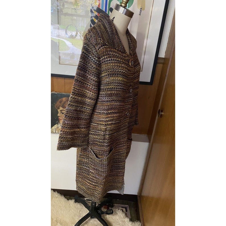 K&ouml;nnte beinhalten: Ein langer, mehrfarbiger Strickmantel mit Schalkragen und zwei Vordertaschen. Der Mantel wird auf einer Schaufensterpuppe pr&auml;sentiert und hat eine Knopfleiste vorne. Die Farbpalette umfasst Braunt&ouml;ne, Gelb und Schwarz.