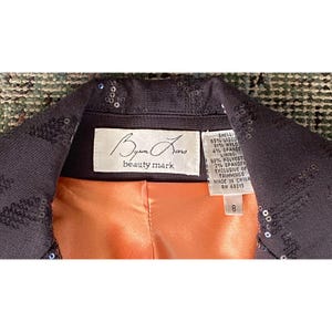 K&ouml;nnte beinhalten: Eine schwarze, mit Pailletten besetzte Jacke mit einem pfirsichfarbenen Futter. Das Etikett lautet "Byun Jano beauty mark" und listet die Stoffzusammensetzung als 65% Viskose, 31% Nylon, 4% Elasthan auf. Das Futter besteht aus 98% Polyester und 2% Elasthan. Die Jacke ist in China hergestellt und hat ein Gr&ouml;&szlig;enetikett mit der Nummer 8.