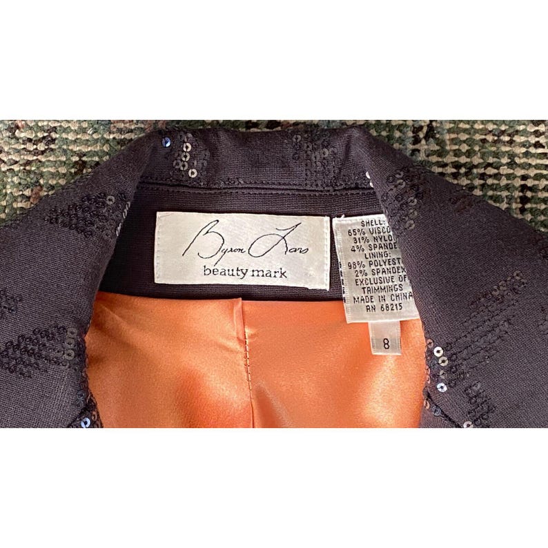 K&ouml;nnte beinhalten: Eine schwarze, mit Pailletten besetzte Jacke mit einem pfirsichfarbenen Futter. Das Etikett lautet "Byun Jano beauty mark" und listet die Stoffzusammensetzung als 65% Viskose, 31% Nylon, 4% Elasthan auf. Das Futter besteht aus 98% Polyester und 2% Elasthan. Die Jacke ist in China hergestellt und hat ein Gr&ouml;&szlig;enetikett mit der Nummer 8.