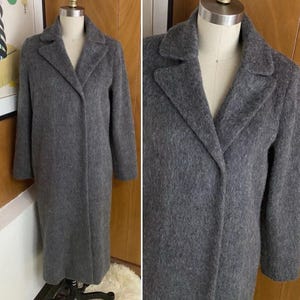 Op de afbeelding: Een lange, grijze jas met een klassieke reverskraag. De jas wordt getoond op een mannequin, waarbij de volledige lengte en de textuur van de stof te zien zijn. Het ontwerp van de jas is eenvoudig en elegant, geschikt voor diverse gelegenheden.