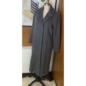 Op de afbeelding: Een lange, grijze wollen jas met een klassieke kraag en een getailleerde pasvorm. De jas wordt gepresenteerd op een mannequin, die de volledige lengte en het ontwerp laat zien. De kleur van de jas is een neutraal grijs, geschikt voor diverse outfits.