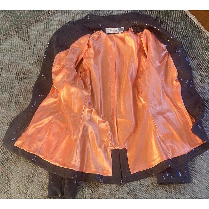 K&ouml;nnte beinhalten: Ein schwarzer Blazer mit Pailletten und einem Futter aus pfirsichfarbenem Satin. Der Blazer hat ein klassisches Design mit einem Schalkragen und langen &Auml;rmeln.