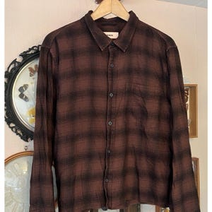 May include: A brown and black checked long-sleeved shirt, buttoned up and hanging on a wooden hanger. The shirt has a collared neckline and a single chest pocket. The fabric appears to be a soft, woven material. The shirt is a classic style.