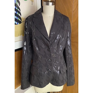 K&ouml;nnte beinhalten: Ein schwarzer Blazer mit einem Pailletten-Hahnentrittmuster. Der Blazer hat einen Schalkragen, lange &Auml;rmel und einen einreihigen Verschluss mit drei Kn&ouml;pfen.