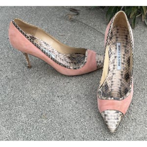 Puede incluir: Un par de tacones altos de ante rosa con un estampado de piel de serpiente en el interior y en la puntera. Los tacones son de estilo stiletto. El nombre de la marca "MANOLO BLAHNIK" es visible en la plantilla.