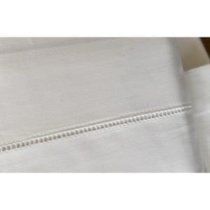 Pu&ograve; includere: Primo piano di tessuto bianco con un design decorativo orizzontale a cuciture aperte. Il tessuto sembra essere un lenzuolo o un altro articolo da letto. La cucitura &egrave; uniformemente distanziata e crea un dettaglio strutturato.