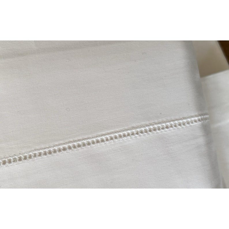 Pu&ograve; includere: Primo piano di tessuto bianco con un design decorativo orizzontale a cuciture aperte. Il tessuto sembra essere un lenzuolo o un altro articolo da letto. La cucitura &egrave; uniformemente distanziata e crea un dettaglio strutturato.