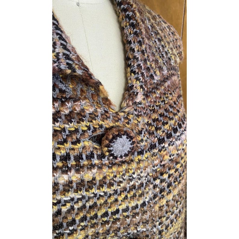 K&ouml;nnte beinhalten: Nahaufnahme eines mehrfarbigen Strickkleidungsst&uuml;cks mit Knopfverschluss. Die Farben sind Braun, Gelb, Grau und Schwarz. Der Knopf ist ein rundes, grau-braunes Design. Das Kleidungsst&uuml;ck wird auf einer Schaufensterpuppe pr&auml;sentiert.