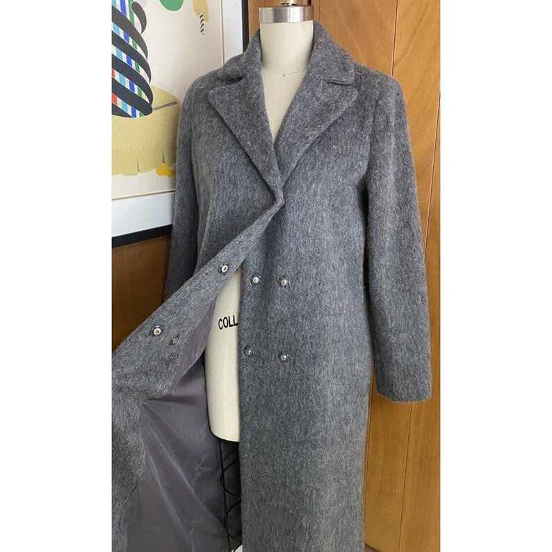 Op de afbeelding: Een lange, grijze wollen jas met een dubbele rij knopen. De jas heeft een klassieke reverskraag, knoopsluitingen en een zacht, getextureerd uiterlijk. De jas wordt op een mannequin gepresenteerd.
