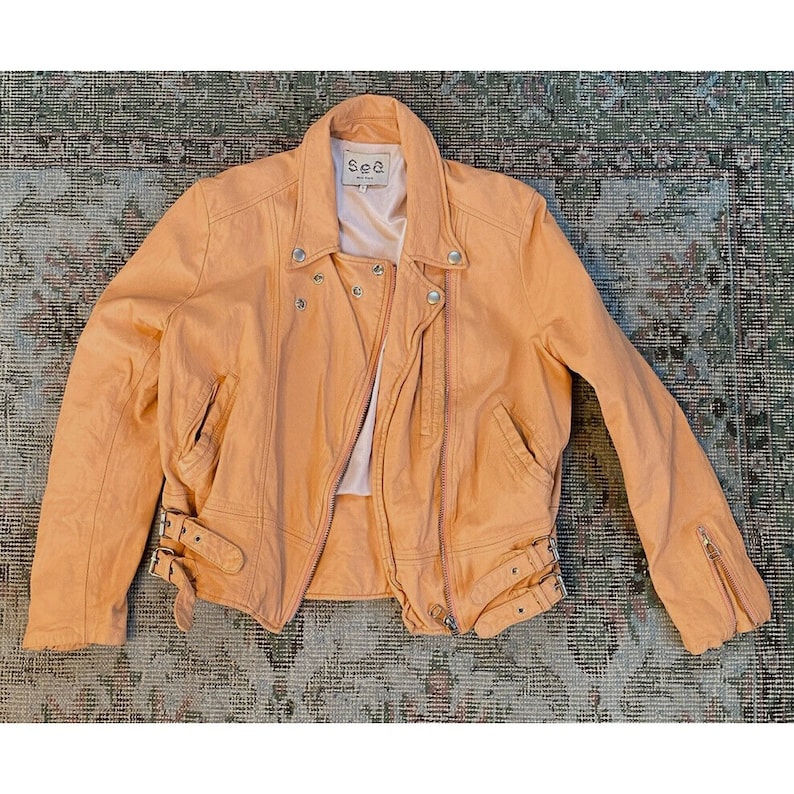 Pode incluir: Uma jaqueta de couro moto de cor laranja claro com fecho de correr e uma etiqueta de marca que diz "Sea" no colarinho.