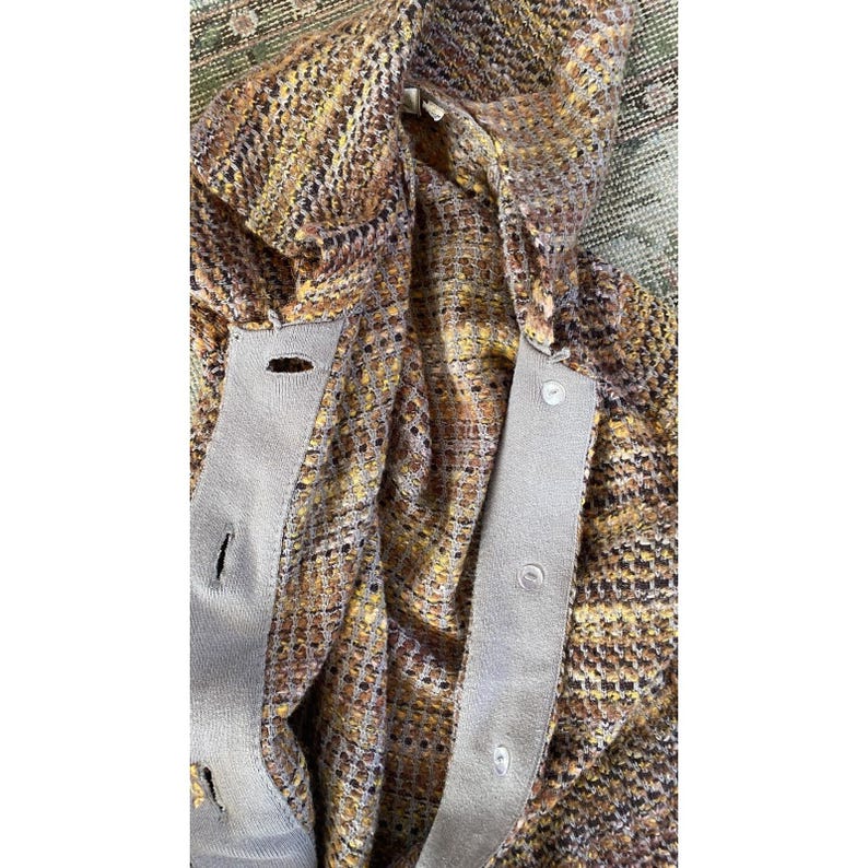 K&ouml;nnte beinhalten: Nahaufnahme einer mehrfarbigen Tweed-Strickjacke mit Knopfleiste. Der Stoff weist eine Mischung aus braunen, gelben und schwarzen F&auml;den auf. Die Strickjacke hat einen hellgrauen Besatz entlang der Knopfleiste und um die Knopfl&ouml;cher.