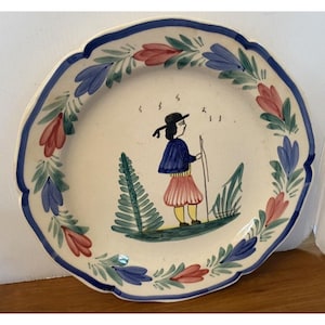 Könnte beinhalten: Ein weißer Keramikteller mit einem blauen und grünen Rand. Die Mitte des Tellers zeigt ein handgemaltes Design einer Person in einem blauen Mantel und einem roten Rock, die einen Stock hält. Die Person steht auf einem grünen Feld mit Bäumen.