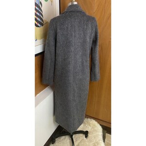 Op de afbeelding: Een lange, grijze wollen jas met een klassieke kraag en een getailleerde pasvorm. De jas wordt getoond op een mannequin, die de volledige lengte en het ontwerp laat zien. De kleur van de jas is een neutraal grijs, geschikt voor diverse outfits.