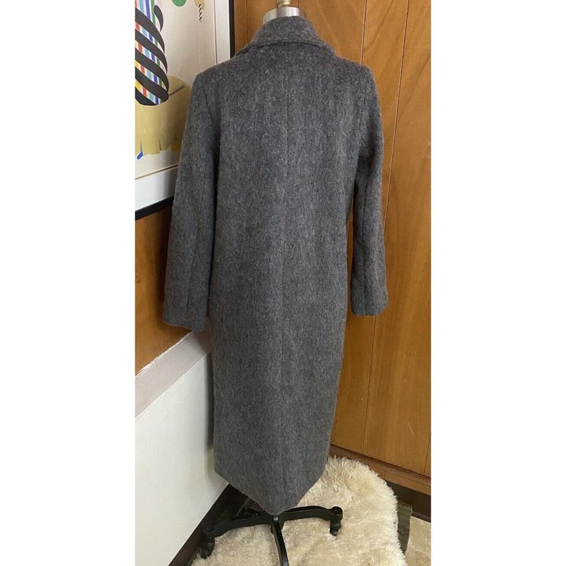 Op de afbeelding: Een lange, grijze wollen jas met een klassieke kraag en een getailleerde pasvorm. De jas wordt getoond op een mannequin, die de volledige lengte en het ontwerp laat zien. De kleur van de jas is een neutraal grijs, geschikt voor diverse outfits.