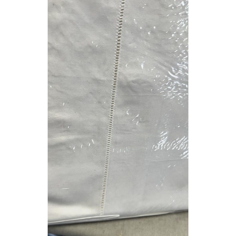 Pu&ograve; includere: Un articolo tessile bianco, forse un lenzuolo o una tovaglia, &egrave; parzialmente visibile. Il tessuto ha una trama sottile e un bordo cucito decorativo che corre verticalmente al centro. L'articolo &egrave; racchiuso in plastica trasparente.