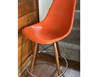 May include: An orange mid-century modern bar stool. The seat is a vibrant orange color, supported by wooden legs and a chrome footrest. The legs are connected by a black metal support structure. The stool is positioned near a wooden door and a carpeted staircase.